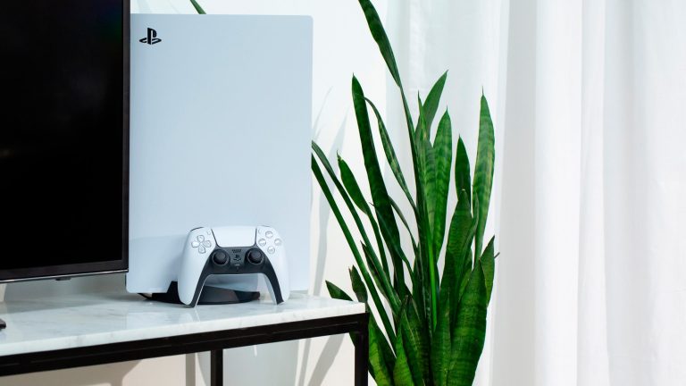 Eine PS5 steht neben einem Fernseher auf einem Marmortisch. Vor der Konsole lehnt der DualSense Controller. Neben dem Tisch ragt eine Pflanze in die Höhe. Eine PS5 steht neben einem Fernseher auf einem Marmortisch. Vor der Konsole lehnt der DualSense Controller. Neben dem Tisch ragt eine Pflanze in die Höhe.