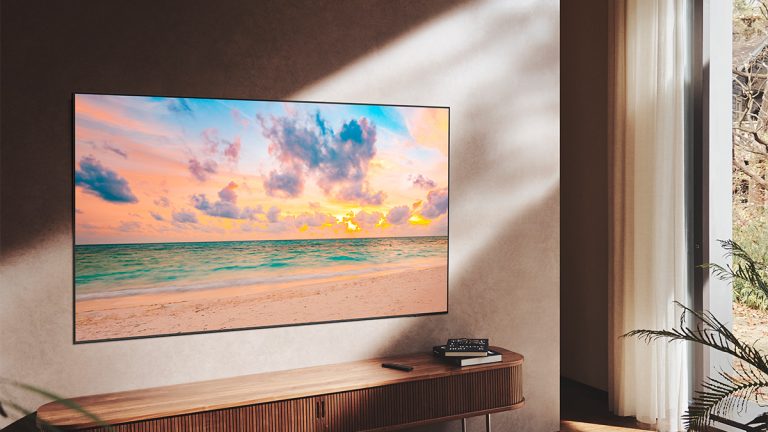 Samsung QN90B Ein QN 90B hängt an einer Wand. Darunter ist eine TV-Bank zu sehen, auf der ein paar Bücher liegen. Auf dem Display ist ein Strand bei Sonnenuntergang dargestellt.