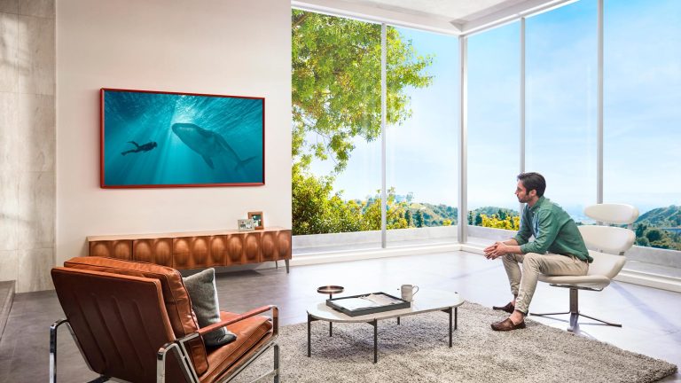 Samsung The Frame Ein Samsung The Frame hängt an einer Wand in einem Wohnzimmer. Eine Person sitzt auf einem Sessel dafür und blickt auf eine Unterwasser-Szene auf dem Bildschirm.