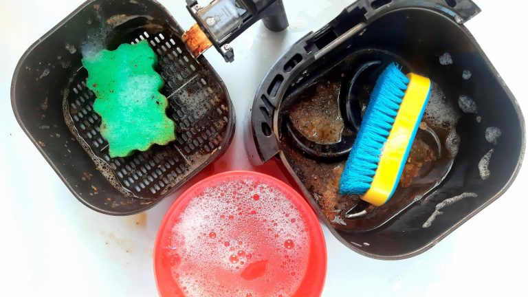 Der Garkorb und die Wanne der Heißluftfritteuse stehen nebeneinander. In beiden Teilen liegen Schwämme. In einem Plastikbehälter befindet sich Wasser mit Spülmittel