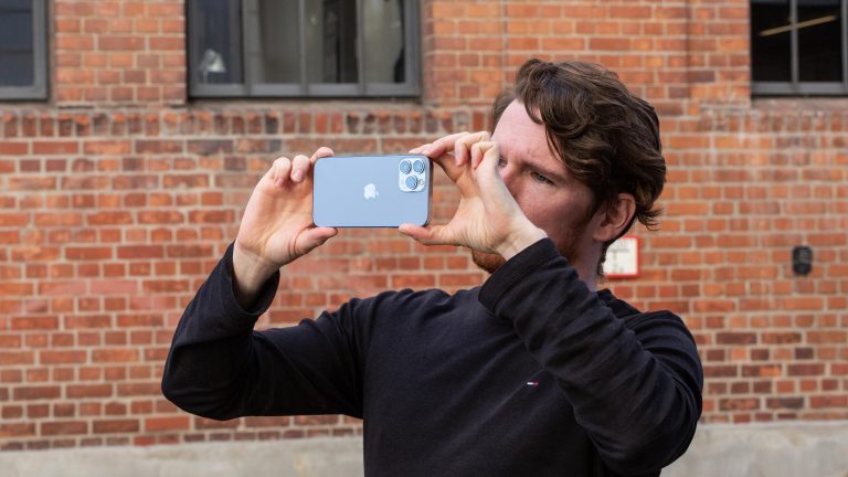 Eine Person schießt mit einem iPhone 13 Pro Max ein Foto. Im Hintergrund ist ein Backsteingebäude zu erkennen.