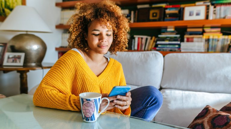 Eine Person sitzt auf einem Sofa und bedient ein Smartphone. Vor ihr steht eine Tasche auf einem Glastisch.