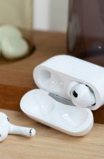 Ein Paar AirPods Pro liegt samt Ladecase auf einem Holzbrett. Einer der beiden Kopfhörer liegt dabei vor der geöffneten Hülle.