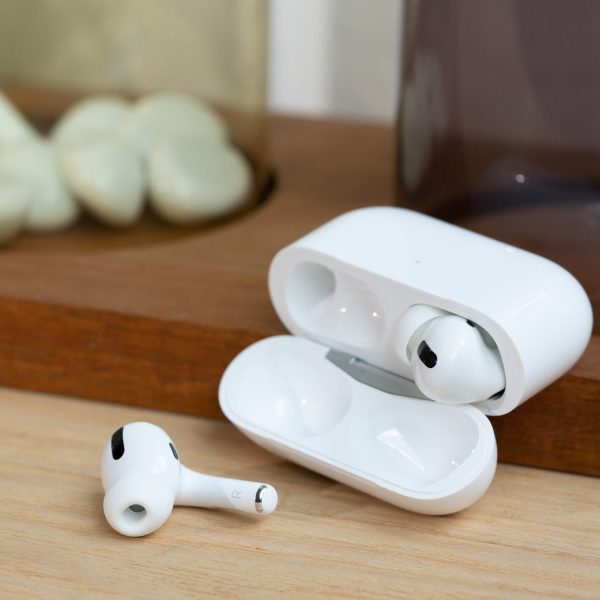 Ein Paar AirPods Pro liegt samt Ladecase auf einem Holzbrett. Einer der beiden Kopfhörer liegt dabei vor der geöffneten Hülle.