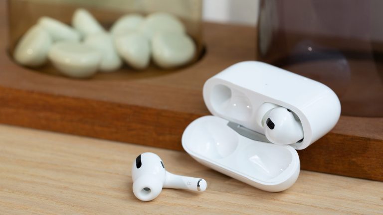 Ein Paar AirPods Pro liegt samt Ladecase auf einem Holzbrett. Einer der beiden Kopfhörer liegt dabei vor der geöffneten Hülle.