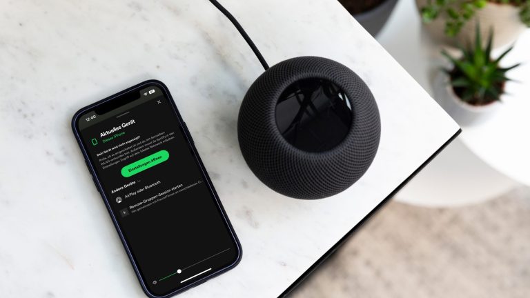 HomePod Spotify Ein schwarzer HomePod steht auf einem Tisch. Daneben liegt ein Smartphone, auf dem die Spotify-App geöffnet wurde.