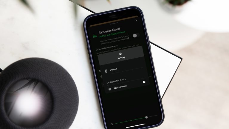 HomePod Spotify Ein schwarzer HomePod steht auf einem Tisch. Daneben liegt ein Smartphone, auf dem die Spotify-App geöffnet wurde.