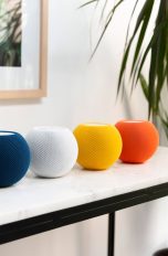 Auf einem Tisch stehen fünf HomePod mini in den Farben Schwarz, Blau, Weiß, Gelb und Orange.