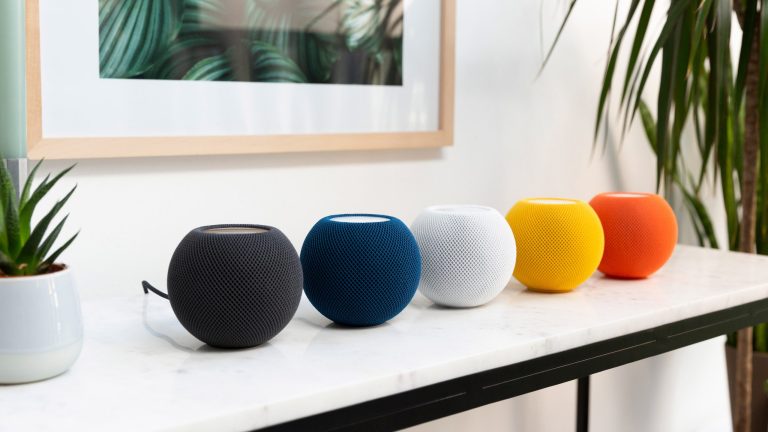 Auf einem Tisch stehen fünf HomePod mini in den Farben Schwarz, Blau, Weiß, Gelb und Orange.