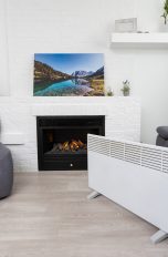 Eine Infrarotheizung steht in einem Wohnzimmer. Im Hintergrund ist ein Karmin zu sehen, auf dem ein Bild steht.