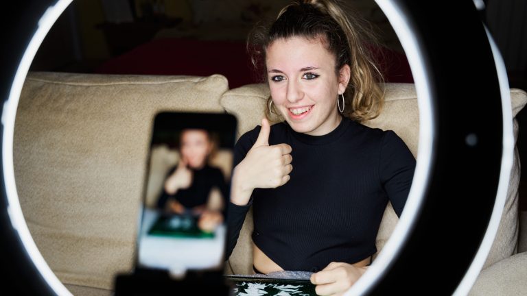 Social Media Stories Eine junge Frau mit blondem Pferdeschwanz sitzt auf einer beigen Couch und hält ihren Daumen in die Smartphone-Kamera.