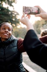 Eine junge Frau tanzt auf offener Straße und lächelt in eine Smartphone-Kamera. Im Vordergrund sieht man Arme, die das Smartphone waagerecht in die Luft halten.