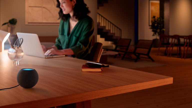 Philips Hue Apple HomeKit Eine Person sitzt an einem Tisch vor einem MacBook. Im Vordergrund steht auf dem Tisch eine HomePod mini.