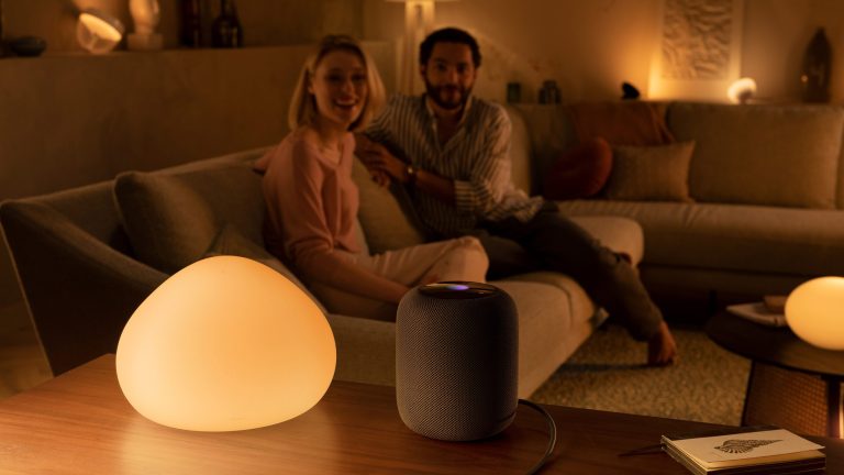 Philips Hue Apple HomeKit Zwei Personen sitzen auf einer Sofalandschaft und blicken in Richtung eines HomePods, der im Vordergrund neben einer Hue-Leuchte steht.