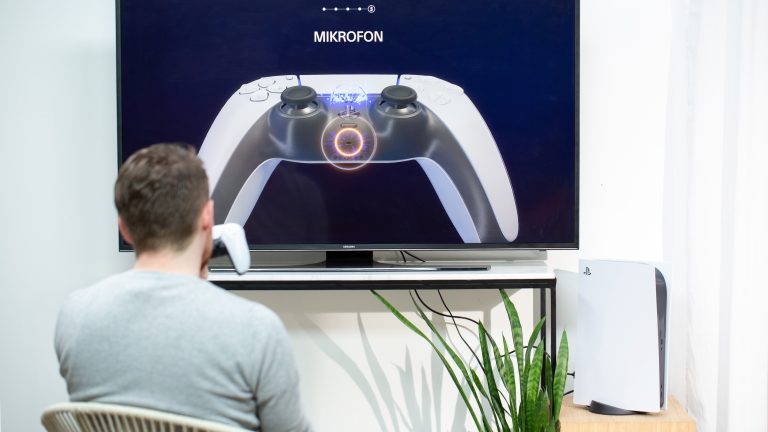 Playstation 5 Dual Sense Eine Person sitzt vor einem Fernseher und spricht ins Mikrofon des DualSense-Controllers.