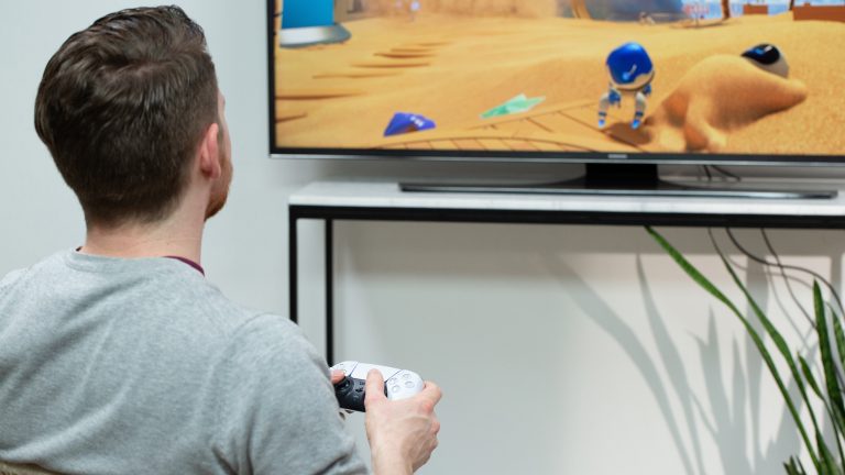 Playstation 5 Dual Sense Eine Person sitzt vor einem Fernseher und hält einen DualSense-Controller in der Hand.