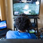 Eine Person sitzt vor einem Fernseher, auf dem gerade ein Computerspiel läuft. Links davon steht ein aufgeklappter Laptop.