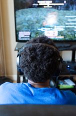 Eine Person sitzt vor einem Fernseher, auf dem gerade ein Computerspiel läuft. Links davon steht ein aufgeklappter Laptop.