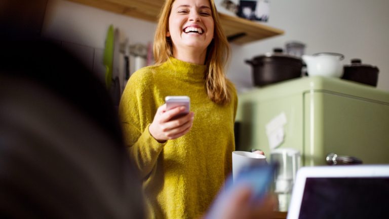 Siri Sprüche Eine lachende Frau steht in einer Küche und hält ein Smartphone in der Hand.