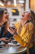 Zwei Personen sitzen in einem Café und lachen ausgelassen. Die eine Person hält ein Smartphone in der Hand.