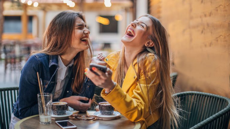 Zwei Personen sitzen in einem Café und lachen ausgelassen. Die eine Person hält ein Smartphone in der Hand.