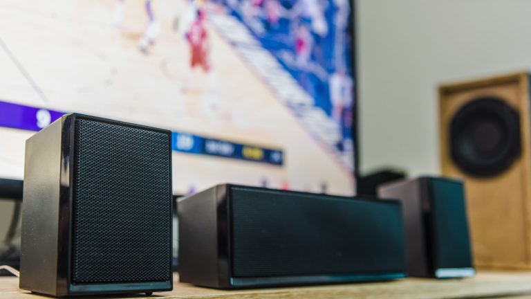 Subwoofer anschließen Drei Lautsprecher stehen vor einem Fernseher in einem Raum. Im Hintergrund ist ein Subwoofer zu sehen.