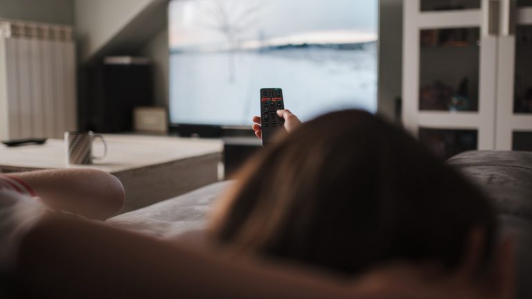 TV Bild einstellen Eine Person liegt auf einem Sofa und hält eine Fernbedienung in die Höhe. Im Hintergrund ist unscharf ein Fernseher zu erkennen, auf dem gerade eine Schneelandschaft gezeigt wird.