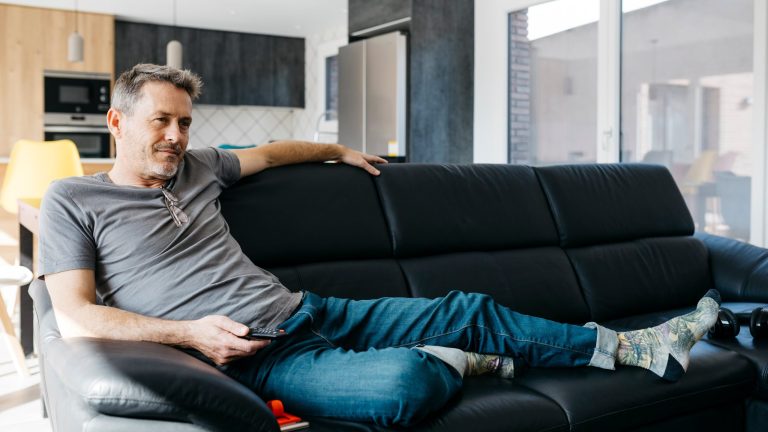 TV Bild einstellen Eine Person liegt entspannt auf einem schwarzen Sofa und hält eine Fernbedienung in der Hand.