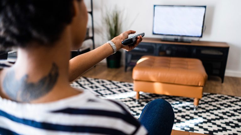 Eine Person sitzt auf einem Sofa und hält eine Fernbedienung Richtung Fernseher.