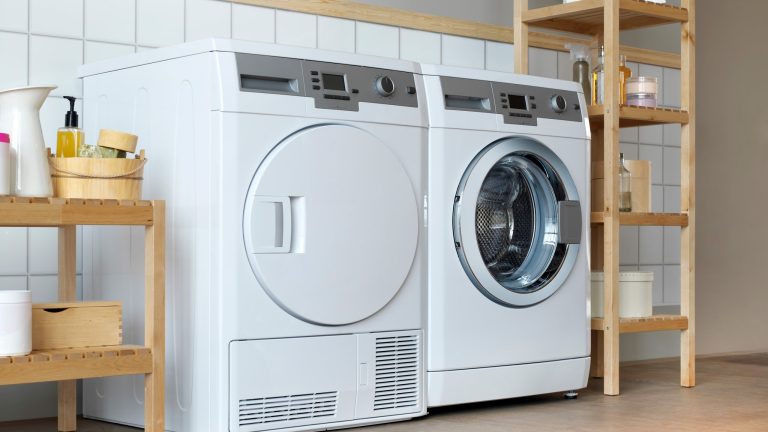 Wäschetrockner reinigen Ein Trockner steht in einem Badezimmer neben einer Waschmaschine. Beide Geräte sind gerahmt von Holzregalen, auf denen diverse Reinigungsmittel stehen.
