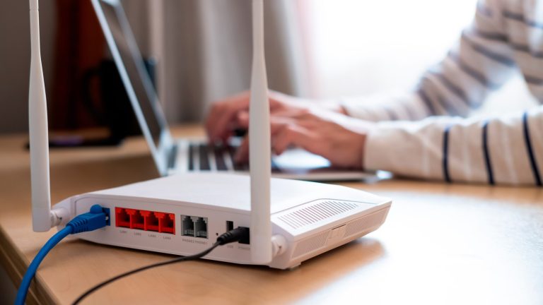 Router einrichten Blick auf die Rückseite eines weißen WLAN-Routers, in den zwei Kabel führen. Dahinter sitzt eine Person und tippt auf einem Laptop.