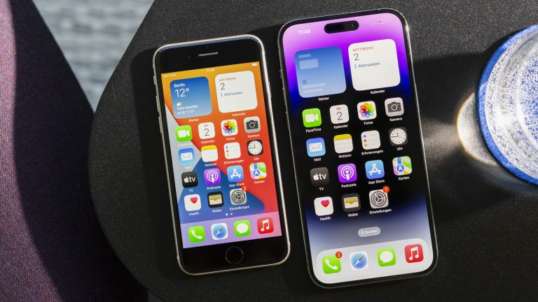 Ein iPhone SE und ein iPhone 14 Pro Max liegen eingeschaltet nebeneinander. Auf beiden Geräten ist der Homebildschirm zu sehen. iPhone 14 Pro Max vs. iPhone SE