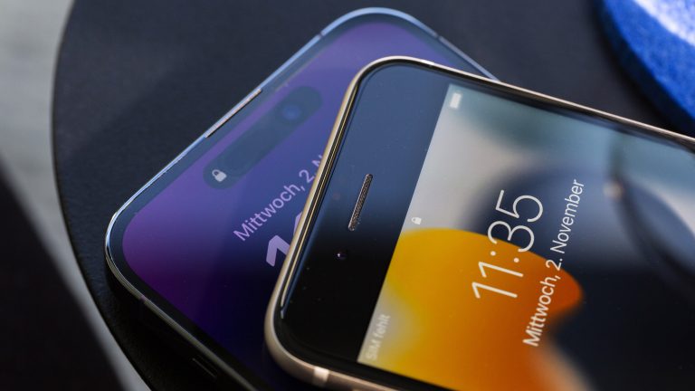Ein iPhone SE und ein iPhone 14 Pro Max liegen aufeinander auf einem schwarzen Tisch. iPhone 14 Pro Max vs. iPhone SE