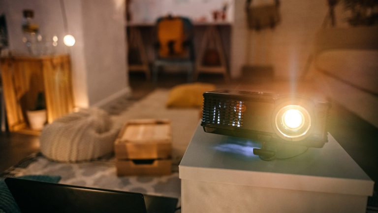 Ein Projektor steht auf einem kleinen Schränkchen in einem Wohnzimmer. Ein Laptop ist an ihn angeschlossen.