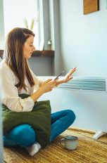 Elektroheizung Eine Person sitzt vor einem Heizlüfter und schaut auf ihr Smartphone.