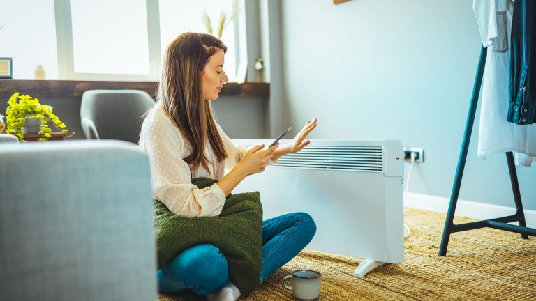 Eine Person sitzt vor einem Heizlüfter und schaut auf ihr Smartphone.