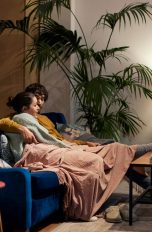 Zwei Personen sitzen auf einem Sofa unter einer Decke und schauen gemeinsam etwas an.