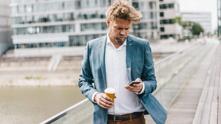 Eine Person mit Kaffeebecher in der Hand spaziert und schaut dabei auf ihr Smartphone.