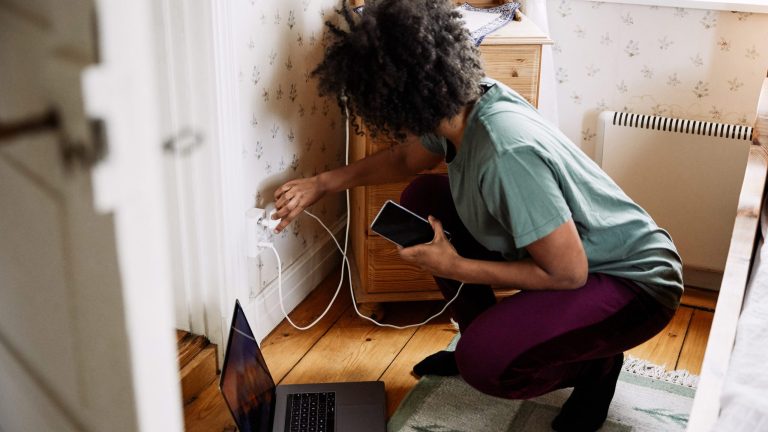 Eine Person kniet vor einer Steckdose, in die sie gerade ein Ladekabel für ihr Smartphone steckt. Eine Person kniet vor einer Steckdose, in die sie gerade ein Ladekabel für ihr Smartphone steckt.