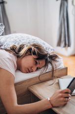 Eine müde aussehende Person liegt im Bett und hält ein Smartphone in der Hand.
