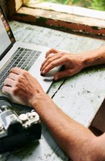 Eine Person sitzt vor einem Laptop und betrachtet Bilder. Daneben liegt eine Kamera.