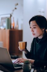 Eine Person mit einer Kaffeetasse in der Hand sitzt vor einem Laptop.