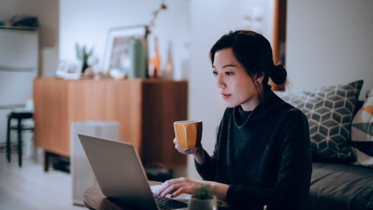 Eine Person mit einer Kaffeetasse in der Hand sitzt vor einem Laptop.
