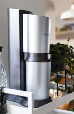 Ein silberner SodaStream steht auf einem weißen Regal neben einer Pflanze und einer Glasflasche.