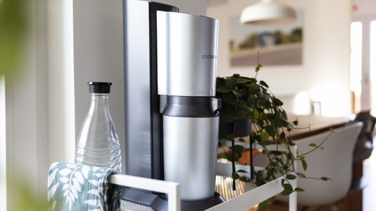 Ein silberner SodaStream steht auf einem weißen Regal neben einer Pflanze und einer Glasflasche.
