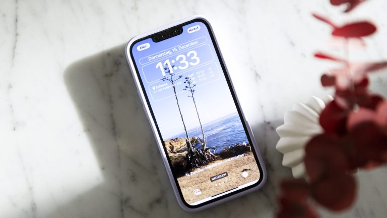iPhone-Hintergrundbild: Festlegen, ändern und anpassen Ein iPhone mit eingeschaltetem Display liegt auf einem Marmortisch. Auf dem Bildschirm ist der neue Sperrbildschirm zu sehen, der sich seit iOS 16 stärker individualisieren lässt.
