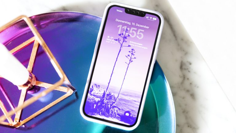 Ein iPhone liegt mit eingeschaltetem Bildschirm in einer Schale. Auf dem Gerät ist ein lilafarbenes Hintergrundbild zu erkennen.