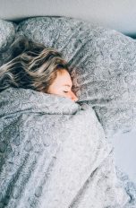 Eine Person liegt bis zur Nase in eine Decke gewickelt in einem Bett. Neben ihr liegt ein Smartphone.