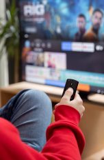 Eine Person sitzt vor einem Fernseher mit der Fernbedienung eines Fire-TV-Sticks in der Hand.