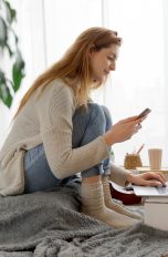 Eine Person sitzt auf einem Sitzkissen vor einem Laptop. In der Hand hält sie ein Smartphone.